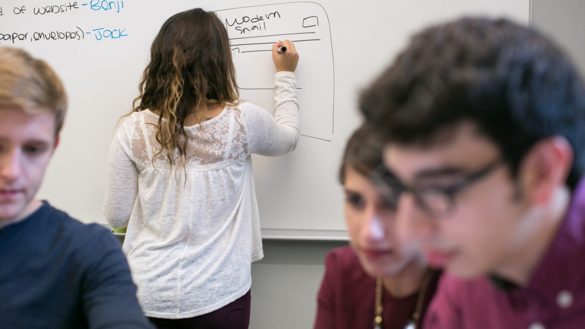 One student sketches on a whiteboard while others sit together in discussion